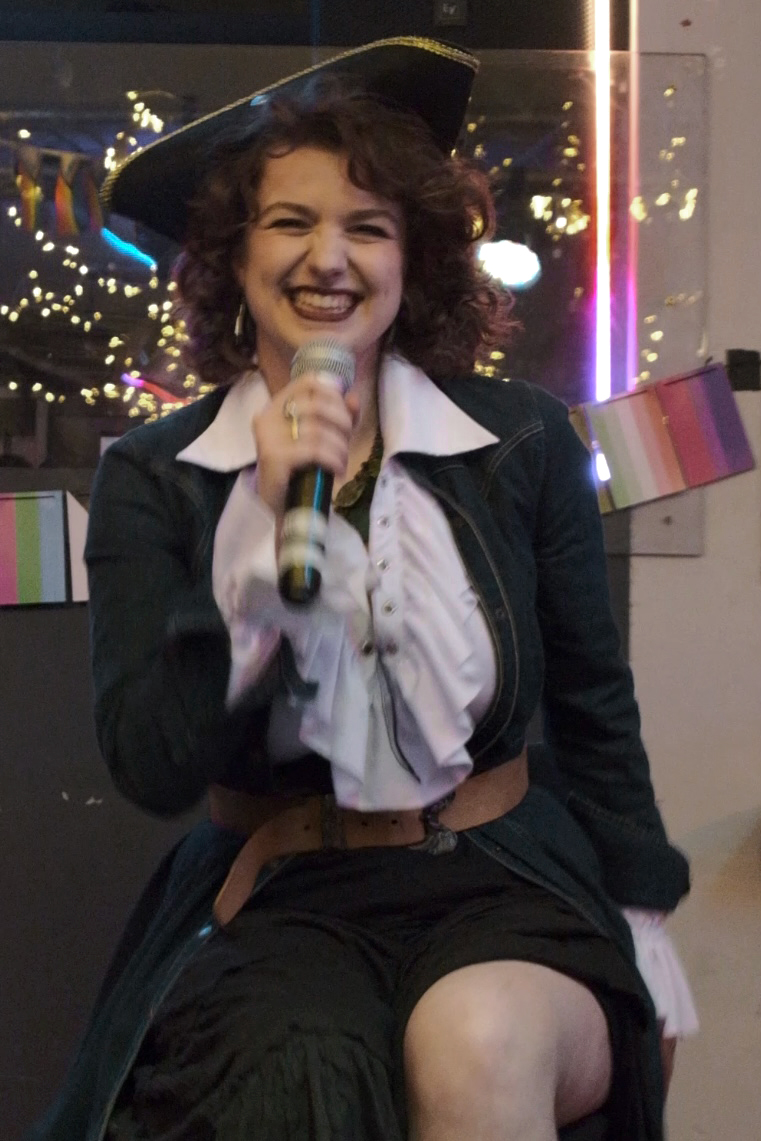 Soph as the pirate Captain Astrid Backwash, holding a microphone and smiling