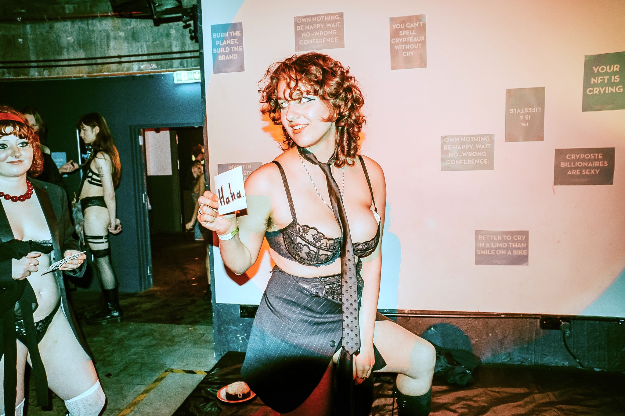 Soph holding a sign that says 'Haha', half dressed in lingerie under a suit that's falling apart